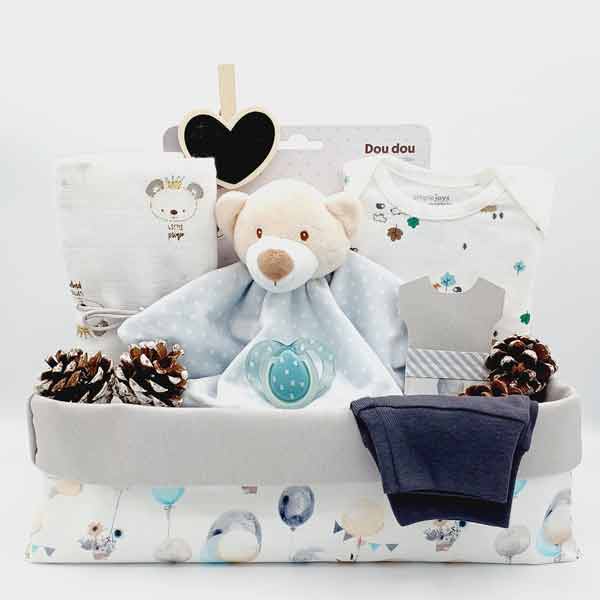 Canastilla de bebé Bosque con doudou de osito azul, piñas decorativas, body, gorrito y pantalón en caja de tela blanca